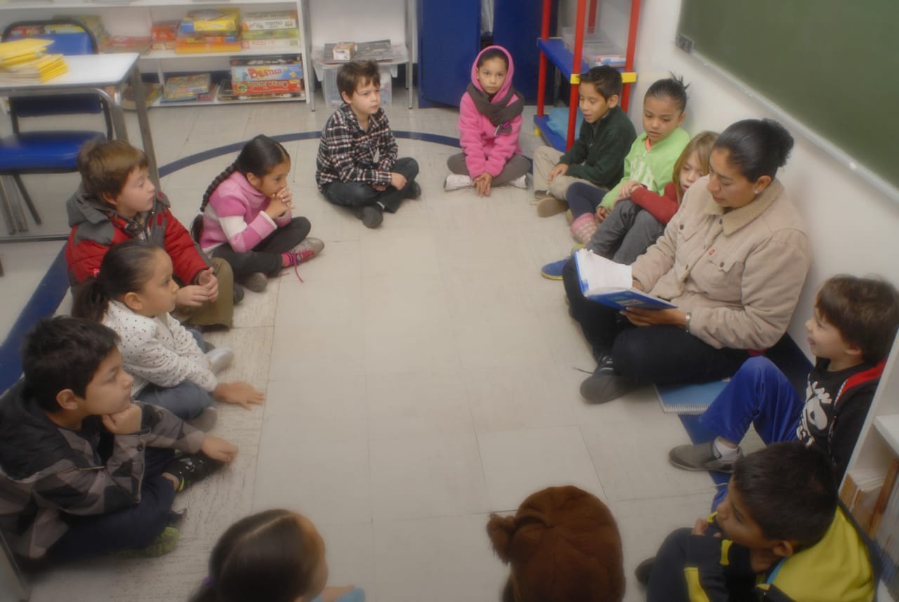 Alumnas y alumnos sentados en círculo escuchan a su maestra leer en el salón