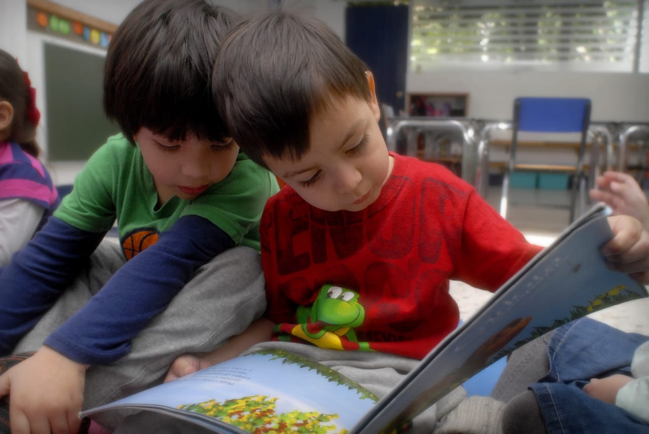 Dos niños leen un libro ilustrado sentados en el suelo del salón