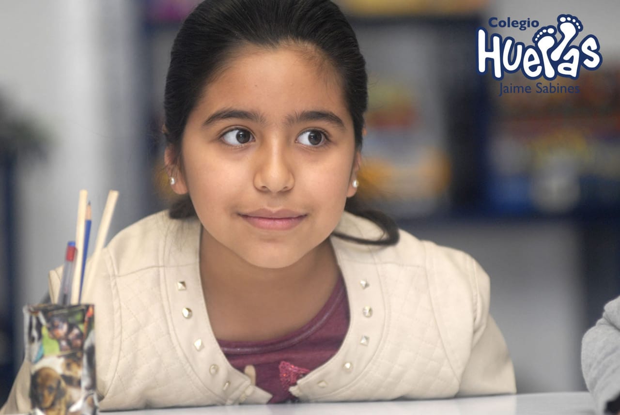 Niña mira de frente con lápices en un vaso sobre su mesa